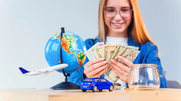 Mulher segurando notas de dinheiro ao lado de um globo, avião em miniatura e carro.