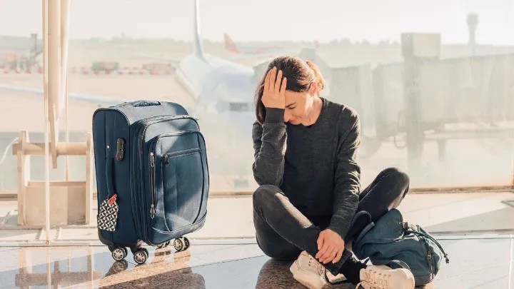 Mulher sentada no chão de um aeroporto ao lado de malas aparentando frustração.