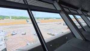 Vista de um saguão interno mostrando a pista de um aeroporto com aviões e veículos no pátio.