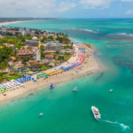 Vista aérea de uma praia com barracas coloridas, mar azul e algumas embarcações próximas.