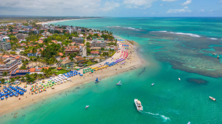 Vista aérea de uma praia com barracas coloridas, mar azul e algumas embarcações próximas.