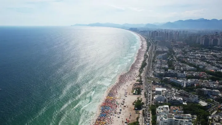 Vista aérea da Praia da Barra da Tijuca com águas claras e faixa de areia cheia.