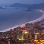 Vista aérea noturna da orla da Barra da Tijuca, com prédios iluminados e o mar ao fundo.