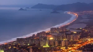 Vista aérea noturna da orla da Barra da Tijuca, com prédios iluminados e o mar ao fundo.