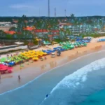 Vista aérea de uma praia com guarda-sóis coloridos, pessoas e mar calmo em Porto de Galinhas.