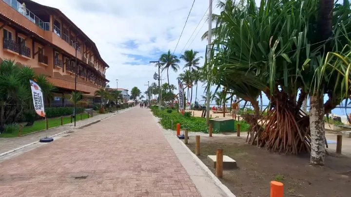 Calçadão com árvores, prédio e vista parcial da Praia do Francês no fundo.
