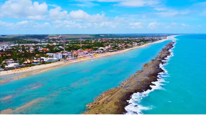 Barra de São Miguel vista de cima com vasta faixa de areia, água azul, muro de corais e prédios ao redor.