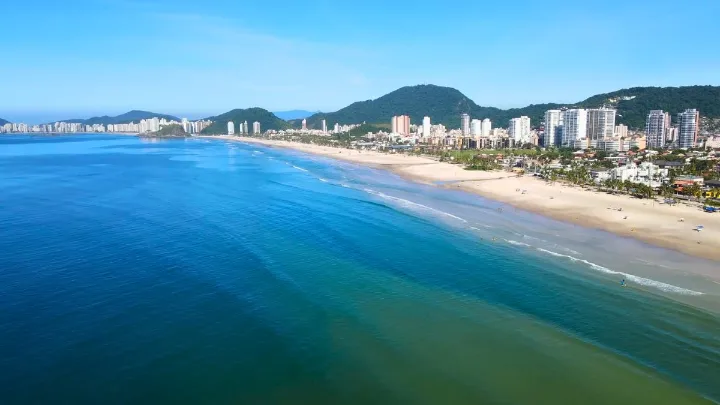 Vista a&eacute;rea da Praia da Enseada Guaruj&aacute; com mar azul, faixa de areia extensa, pr&eacute;dios &agrave; beira-mar e morros ao fundo.