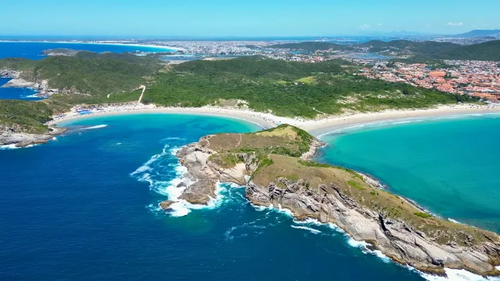 Vista a&eacute;rea da Praia do Forte Cabo Frio com mar azul, faixa de areia clara, cost&otilde;es rochosos, vegeta&ccedil;&atilde;o e &aacute;rea urbana ao fundo.