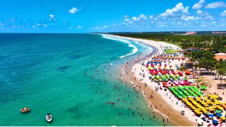 Vista de cima da Praia do Franc&ecirc;s com vasta faixa de areia, banhistas, barracas coloridas, barcos na &aacute;gua e palmeiras ao fundo.