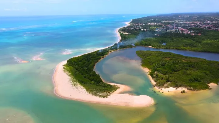 Vista a&eacute;rea da Praia do Patacho com mar claro, faixa de areia, vegeta&ccedil;&atilde;o, lagoa e &aacute;rea urbana ao fundo.