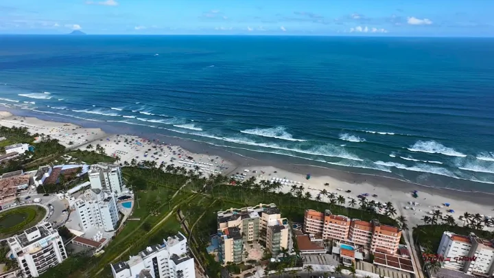 Praia da Riviera de São Lourenço vista de cima, com ampla faixa de areia, vários banhistas, hotéis, prédios e coqueiros ao redor.